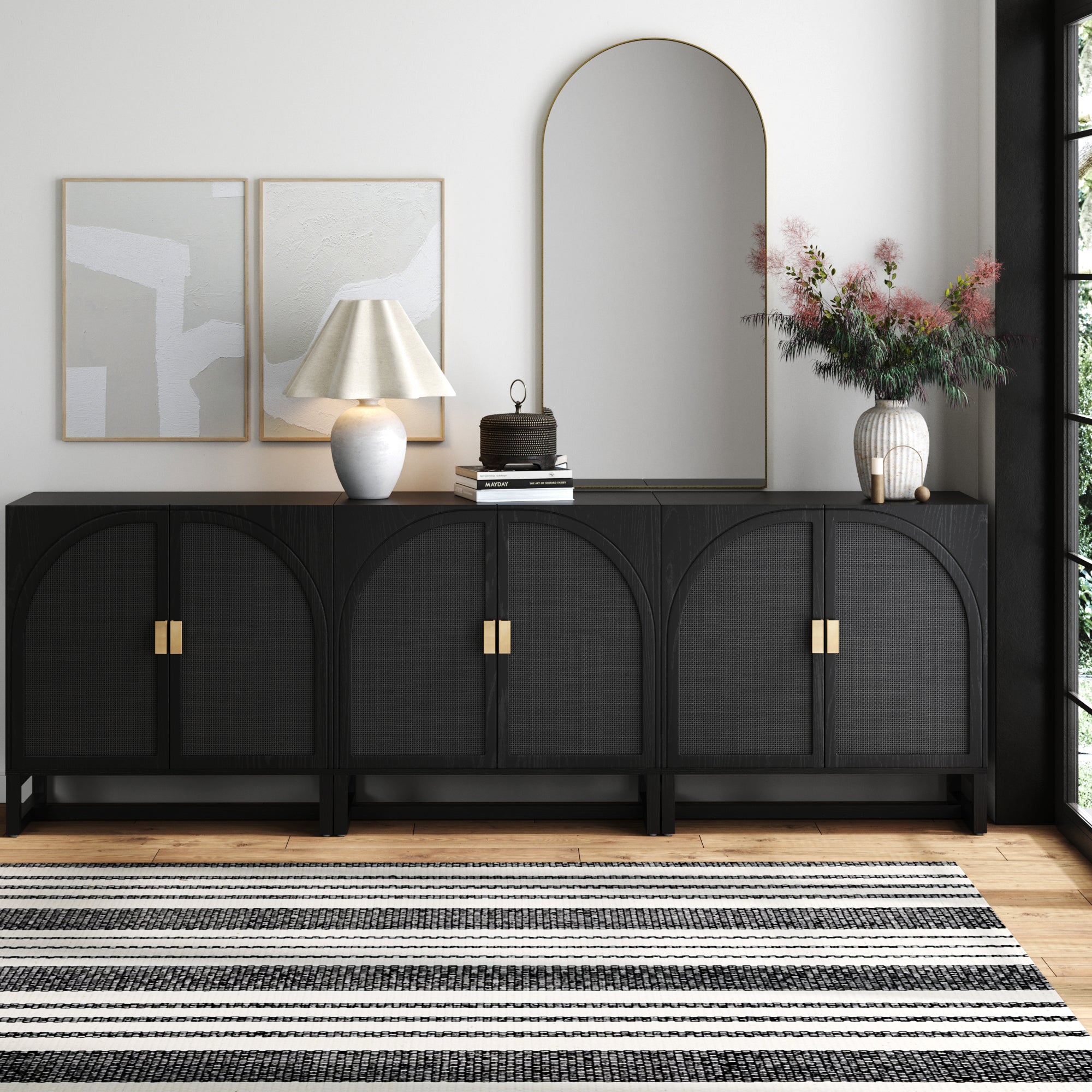A modern living room features the Nathan James Arched Wooden TV Console Credenza with black rattan, gold handles, a large arched mirror, abstract wall art, a white sculptural lamp, decorative boxes, a striped rug, and greenery in a vase.
