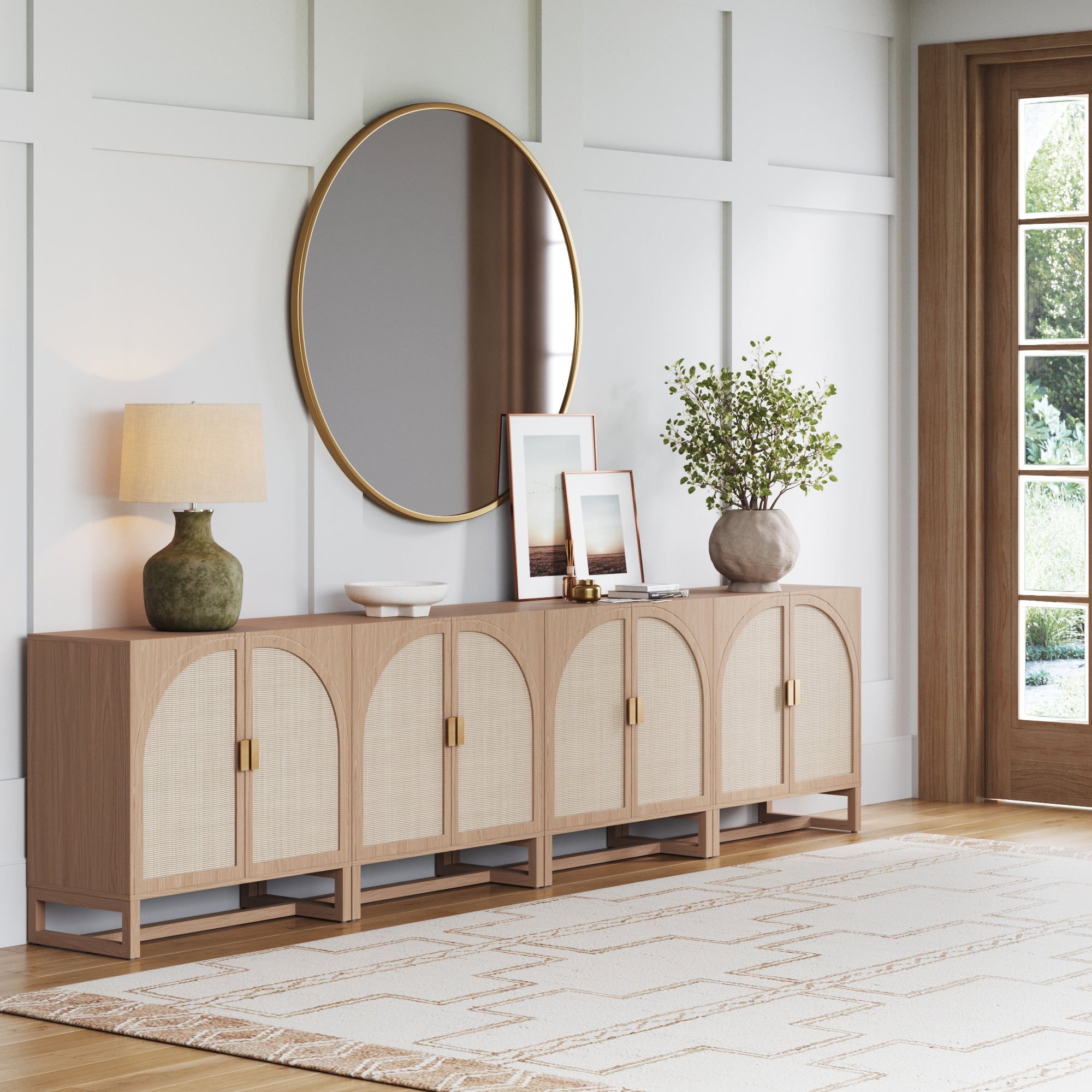 A modern living room with a light wood sideboard, decorative items, a lamp, framed photos, a round mirror with a gold frame, a potted plant, and a patterned rug on a light wood floor.