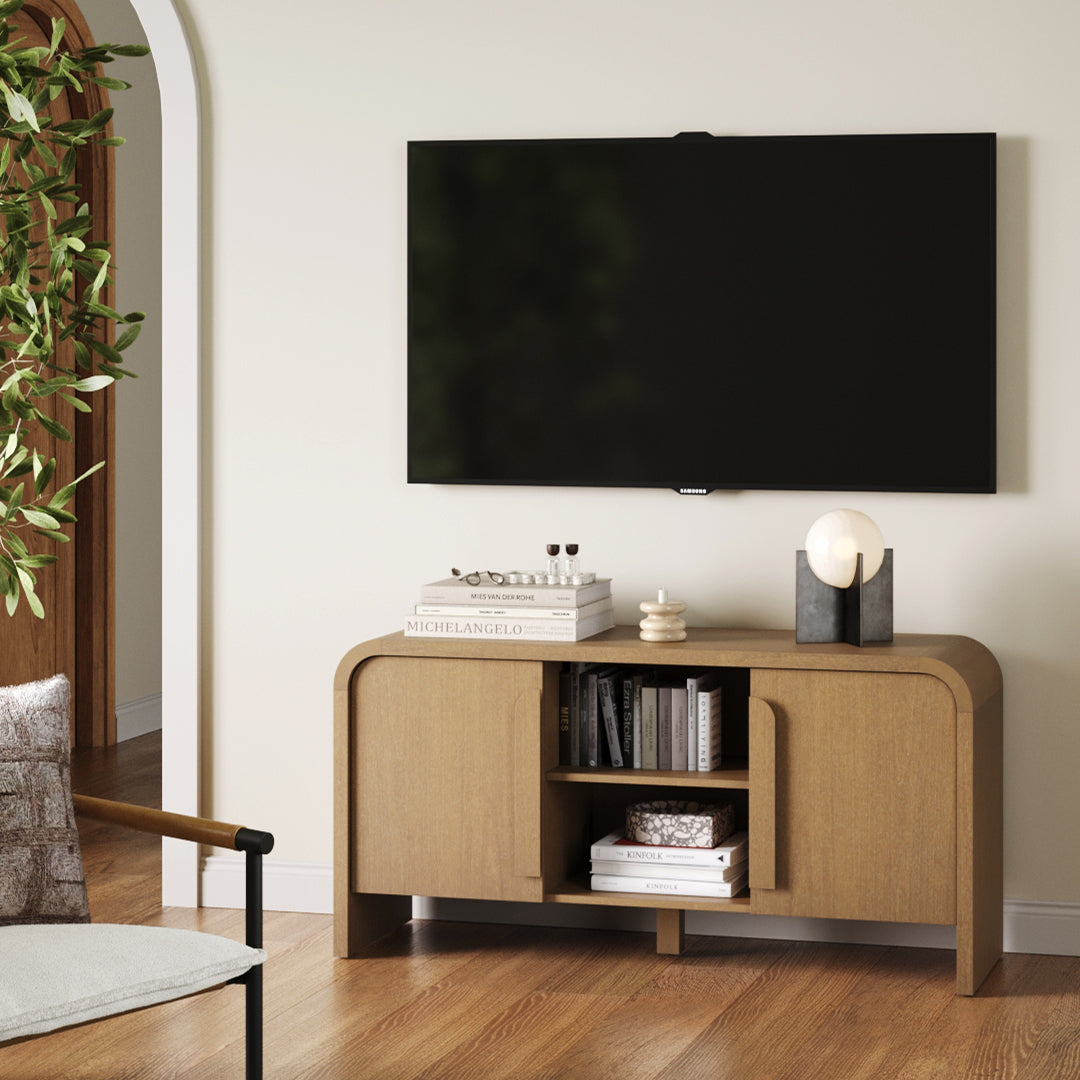 A modern living room with a wall-mounted flat-screen TV above a wooden console table, which holds books, a small sculpture, a candle, and a decorative lamp. A chair and a potted plant are partially visible on the left.