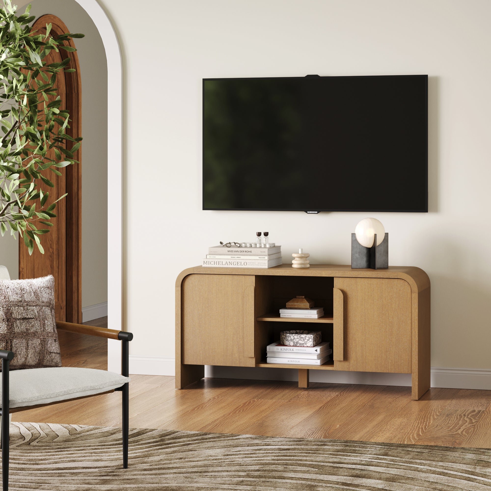 A modern living room features the Nathan James Arched TV Stand or Media Console Cabinet in light brown beneath a wall-mounted TV, topped with books, decor, and a lamp. A green plant and part of a chair are visible on the left.