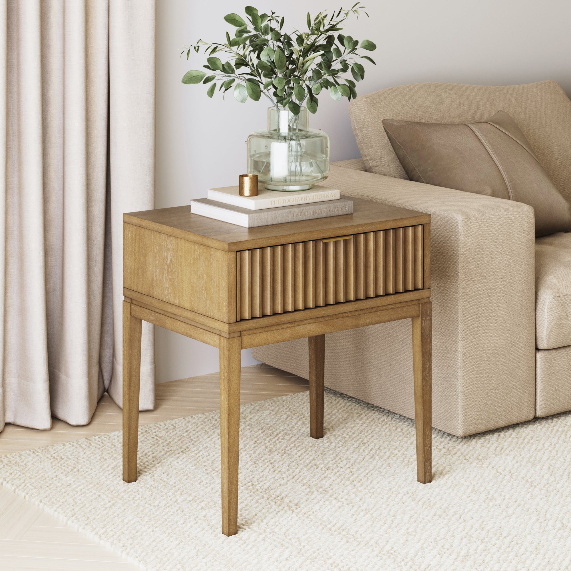 A modern wooden side table with vertical grooves stands next to a beige sofa. On the table are books, a candle, and a glass vase with leafy greenery. A cream curtain and textured rug complete the cozy setting.