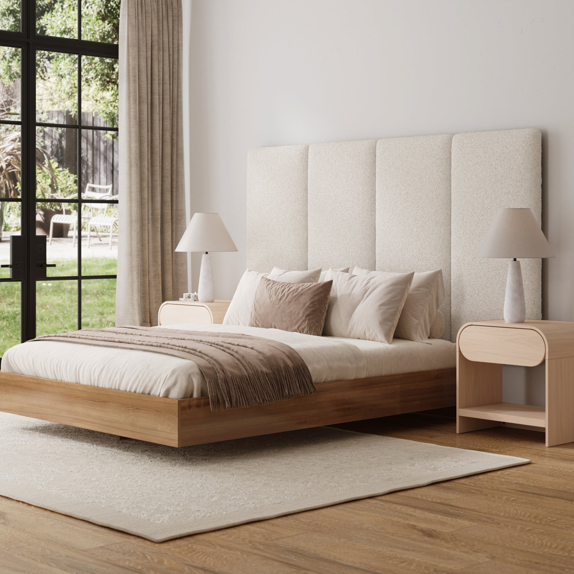 A modern bedroom featuring the Nathan James Wall Mounted 4 Panel Boucle Headboard for King Bed, a wooden platform bed with beige bedding, two matching nightstands with white lamps, a light rug on wooden floors, and a window overlooking greenery.