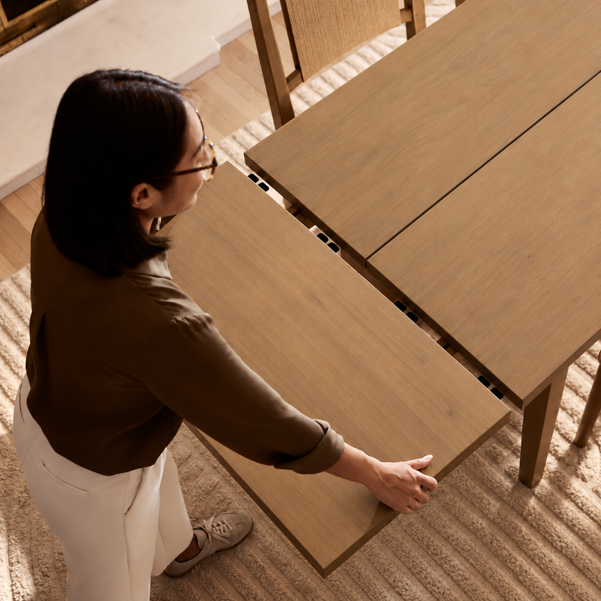 A woman stands at a wooden dining table, pulling a leaf extension into place to lengthen the table.